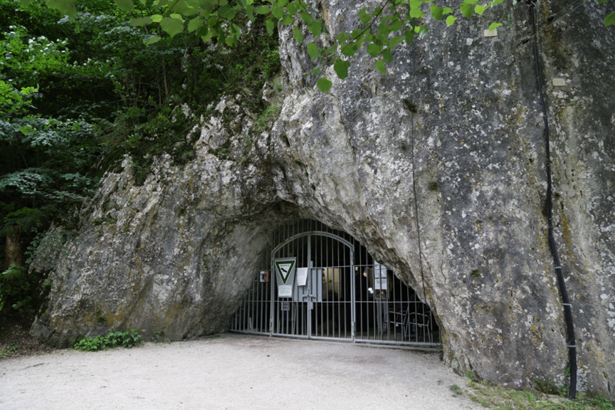 Eine hohe Felswand mit einer großen Eingangsöffnung in die Höhle „Hohler Fels“. Der Eingang ist vergittert. 